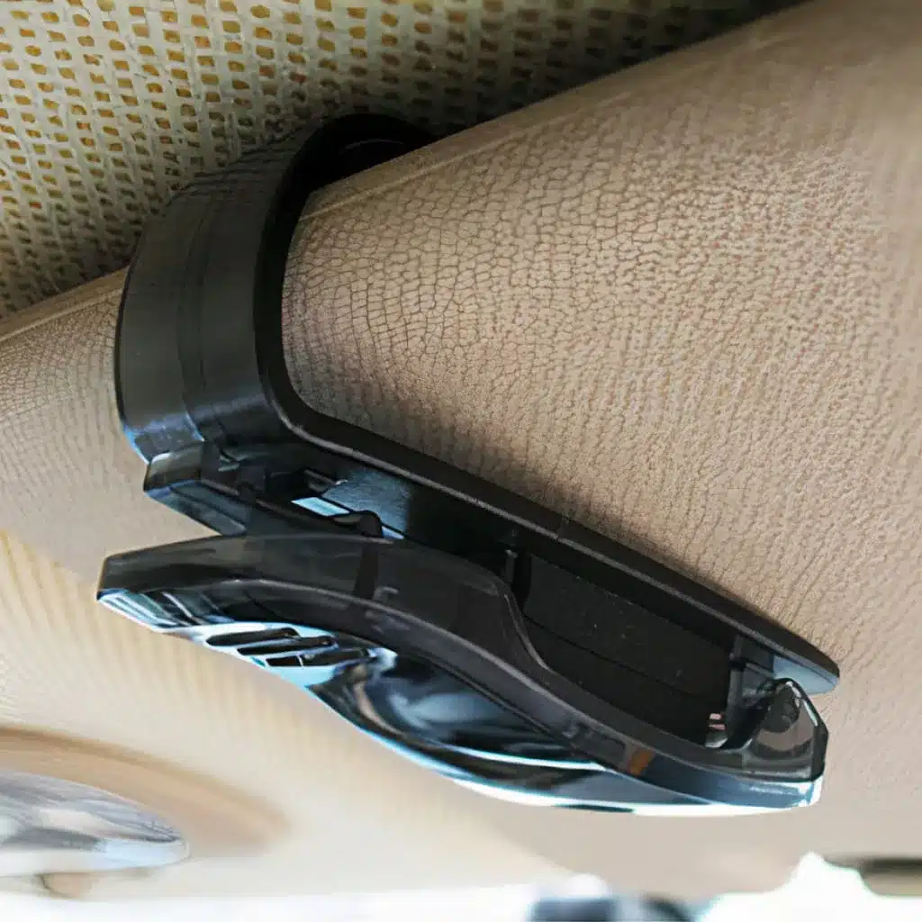 Close-up of a black plastic clip attached to a textured beige surface, likely the interior of a vehicle, designed to hold sunglasses or other small items.