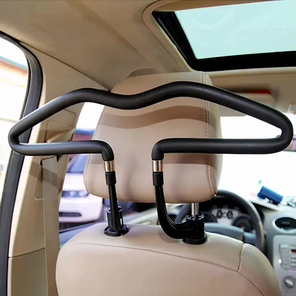 A black car seat coat hanger attached to the back of a beige car seat headrest inside a vehicle. The hanger is designed for hanging clothes and is positioned in front of a sunroof. The steering wheel is visible in the background.