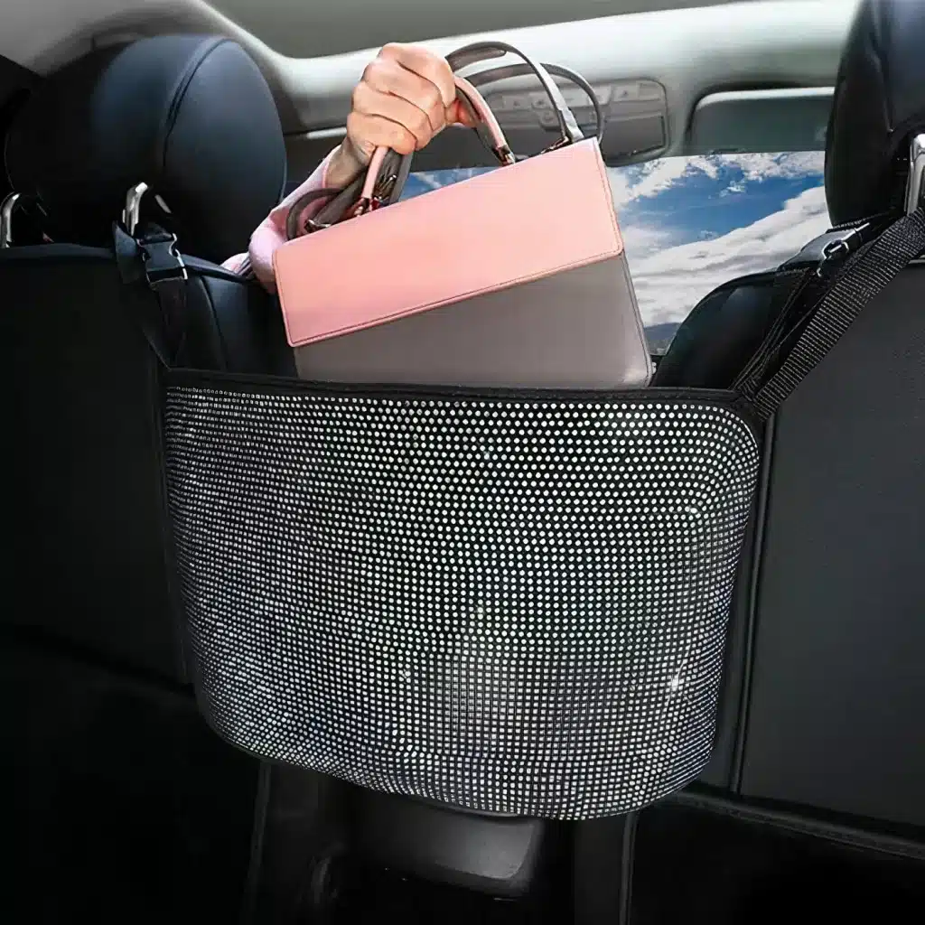 A person places a pink and brown handbag into a net pocket between the front seats of a car. The car seats are black, and a cloudy sky is visible through the back window.