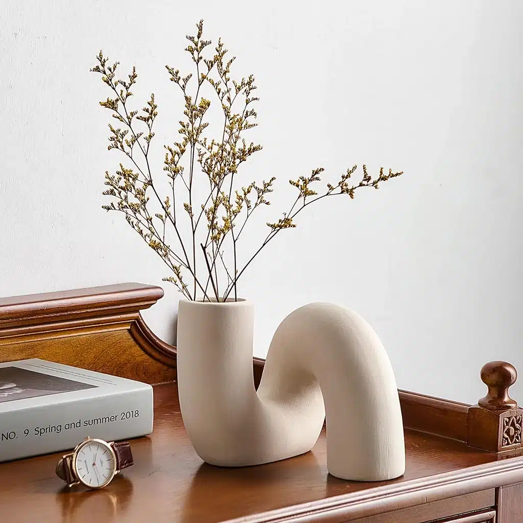 A uniquely shaped beige vase with dry flowers on a wooden surface next to a book and a wristwatch.