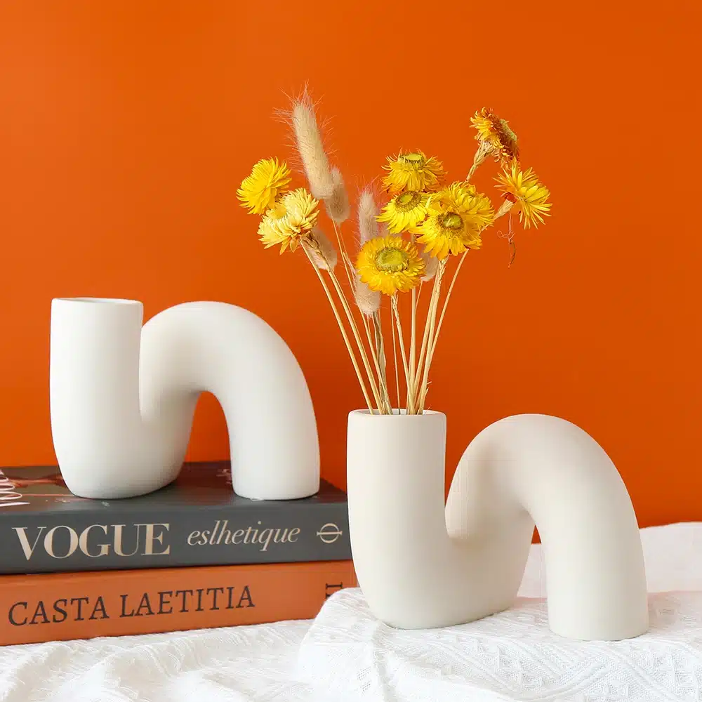 A white, abstract, U-shaped vase holds yellow flowers on a white surface. Next to it is a similar vase without flowers. Two books, Vogue and Casta Laetitia, rest underneath, with an orange background.