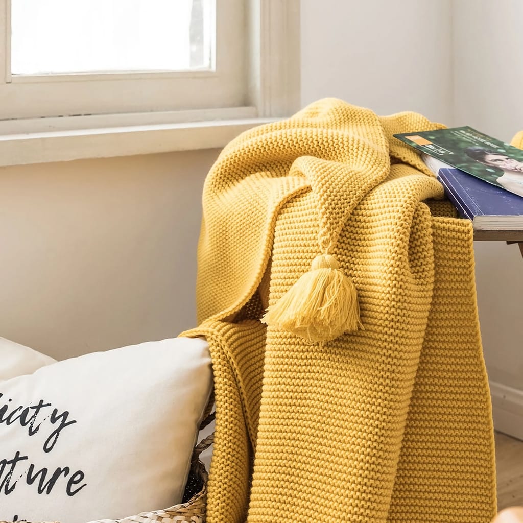 A cozy corner with a folded mustard yellow knitted blanket and a pillow with cursive text. A book with a blue cover rests on the soft surface. Sunlight filters through a window, casting a warm, inviting glow over the scene.