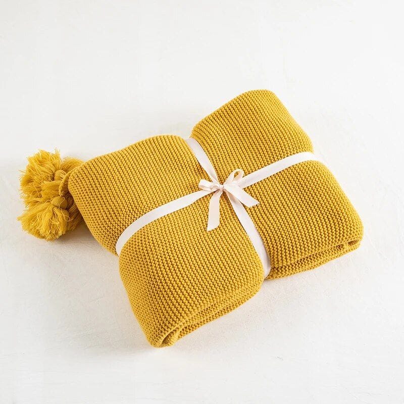 A neatly folded mustard yellow knitted blanket tied with a white ribbon, featuring tassels on one end, placed on a white surface.