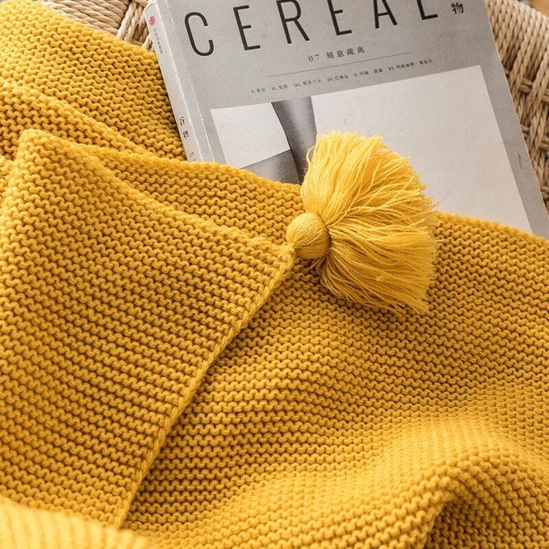 A cozy, mustard-colored knitted blanket with a tassel is draped over the corner of a chair. A magazine titled Cereal with a monochrome cover peeks out from underneath the blanket.