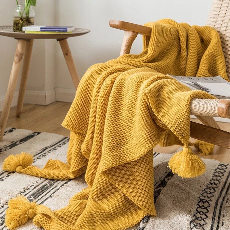 A cozy, mustard-yellow knit blanket with tassels is draped over a woven chair. A round wooden side table holds a book and a plant. The room features light wood flooring and a patterned rug in neutral tones.