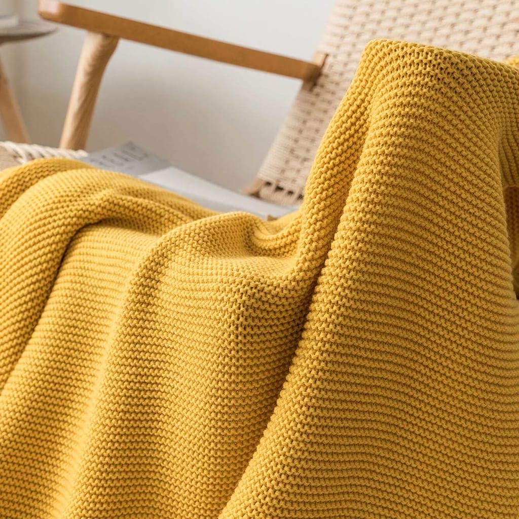 A knitted yellow blanket draped over a woven chair, with a soft and cozy texture. The background shows part of a wooden chair and a white wall, creating a warm and inviting atmosphere.