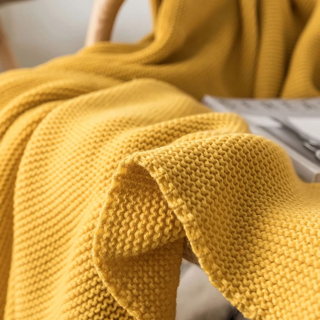 Close-up of a textured, mustard-yellow knit blanket draped over a wooden chair. A magazine lies partially visible on the chair, adding a cozy, relaxed atmosphere to the scene.