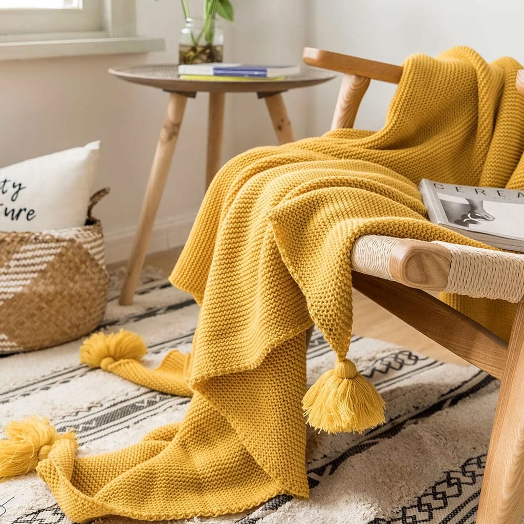 A cozy living room scene with a mustard yellow knitted blanket draped over a wooden chair. A magazine rests on the chair, and a woven basket is nearby. A small wooden table by the window holds a plant and books, and a patterned rug lies beneath.