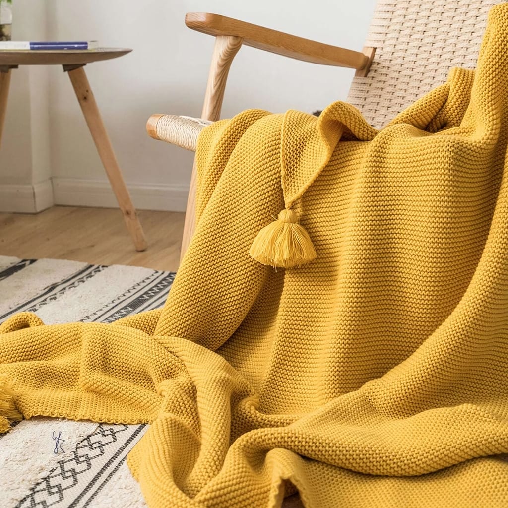 A cozy yellow knitted blanket with a tassel draped over a wicker chair. The chair sits on a light patterned rug, and a small wooden table is in the background. The room has light-colored walls and a wooden floor.