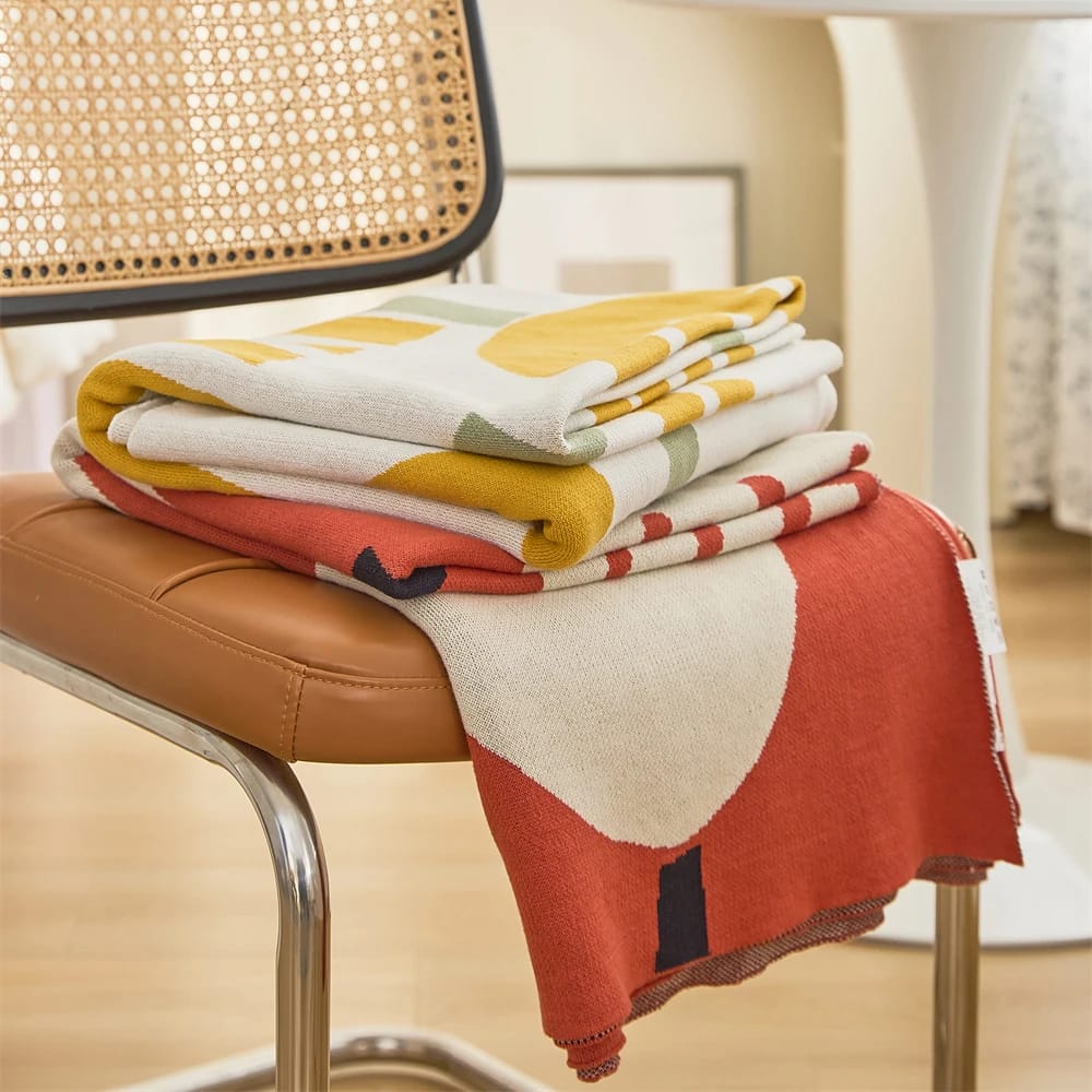 A folded, colorful knitted blanket with abstract patterns sits on a brown chair with a woven backrest. The blanket features shades of red, yellow, and green. A white round table is partially visible in the background on a wooden floor.