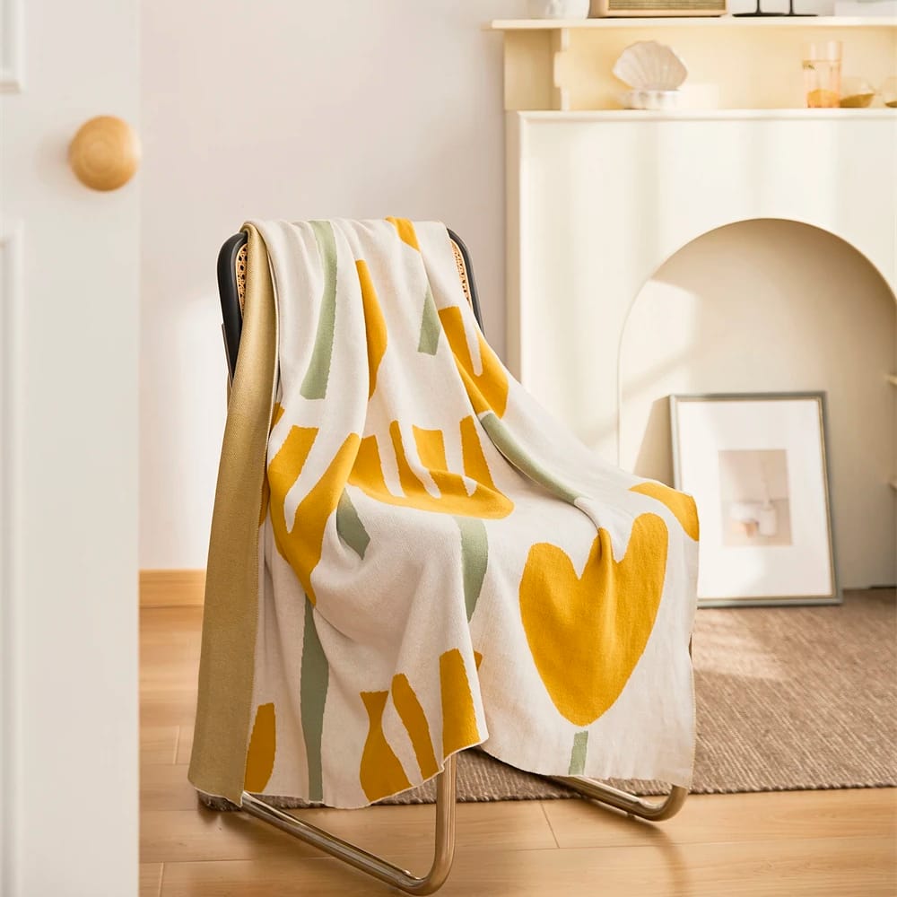 A cozy room with a modern chair draped in a white blanket featuring large yellow and green abstract shapes. The background includes a minimalist shelf with small decor items, a framed picture on the floor, and soft natural lighting.