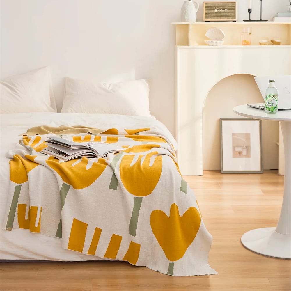 A cozy bedroom with a white bed, draped with a blanket featuring yellow and green abstract shapes. A round white table holds a green glass bottle, and a minimal shelf displays decor, including a Marshall speaker. Soft natural light fills the room.