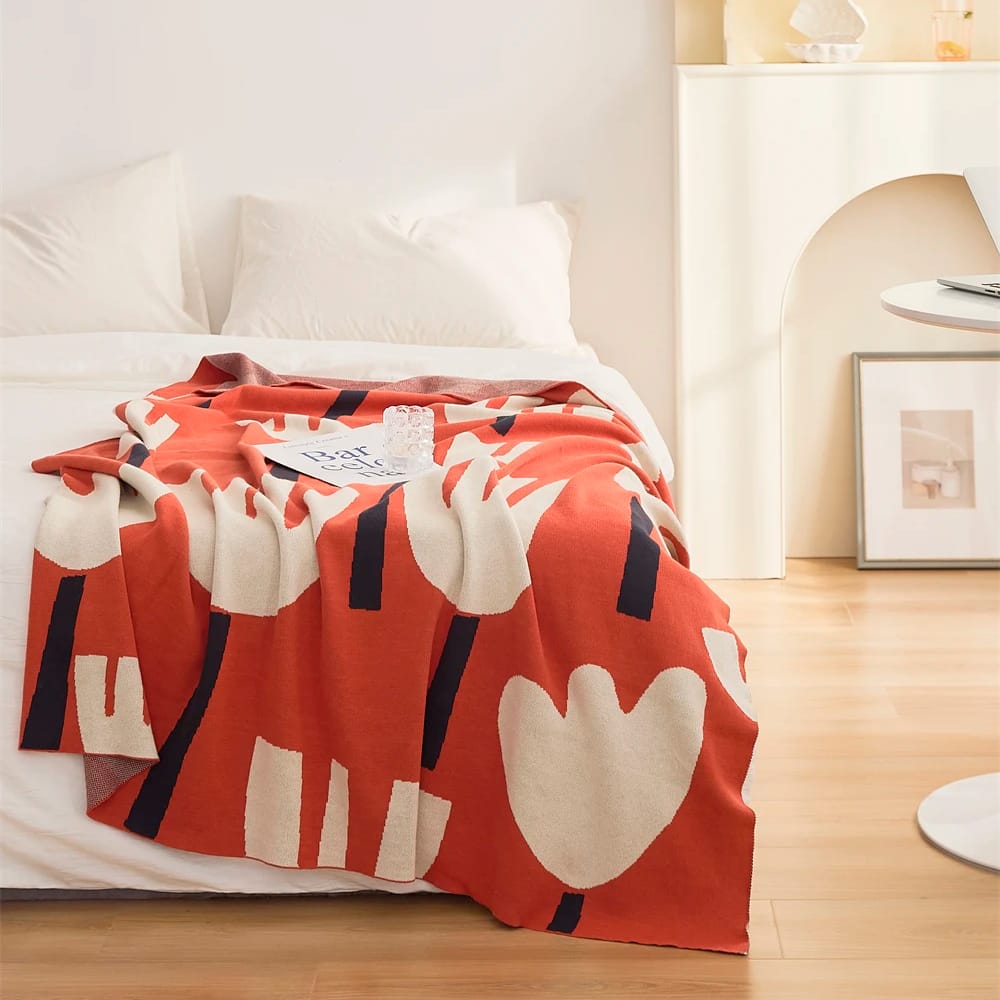 A cozy bedroom with a bed featuring a red blanket with a cream abstract design. The bed is neatly made with white pillows, and a small book lies on the blanket. Soft, warm lighting enhances the rooms inviting atmosphere.