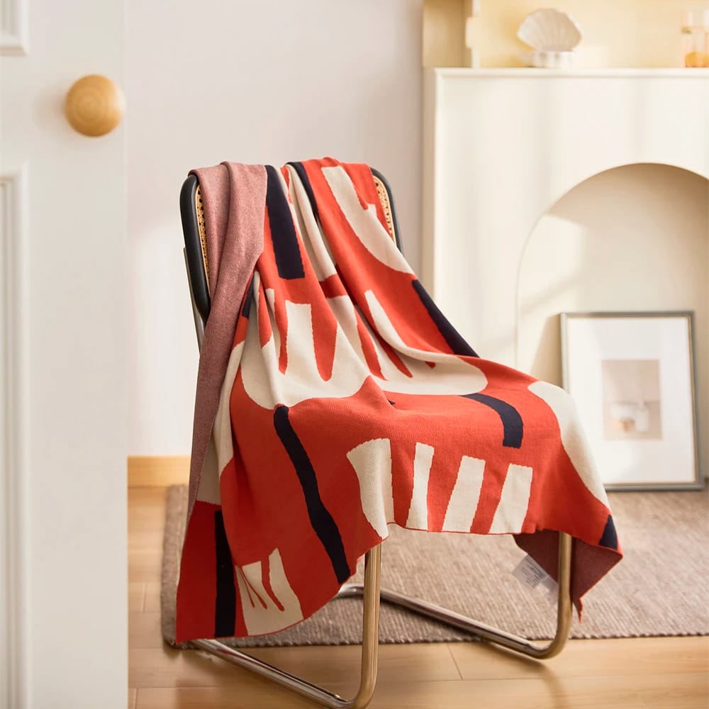 A modern chair draped with a colorful blanket featuring abstract patterns in red, white, and black. The setting is a cozy room with wooden floors and a soft rug, a white fireplace, and a framed picture leaning against the wall.