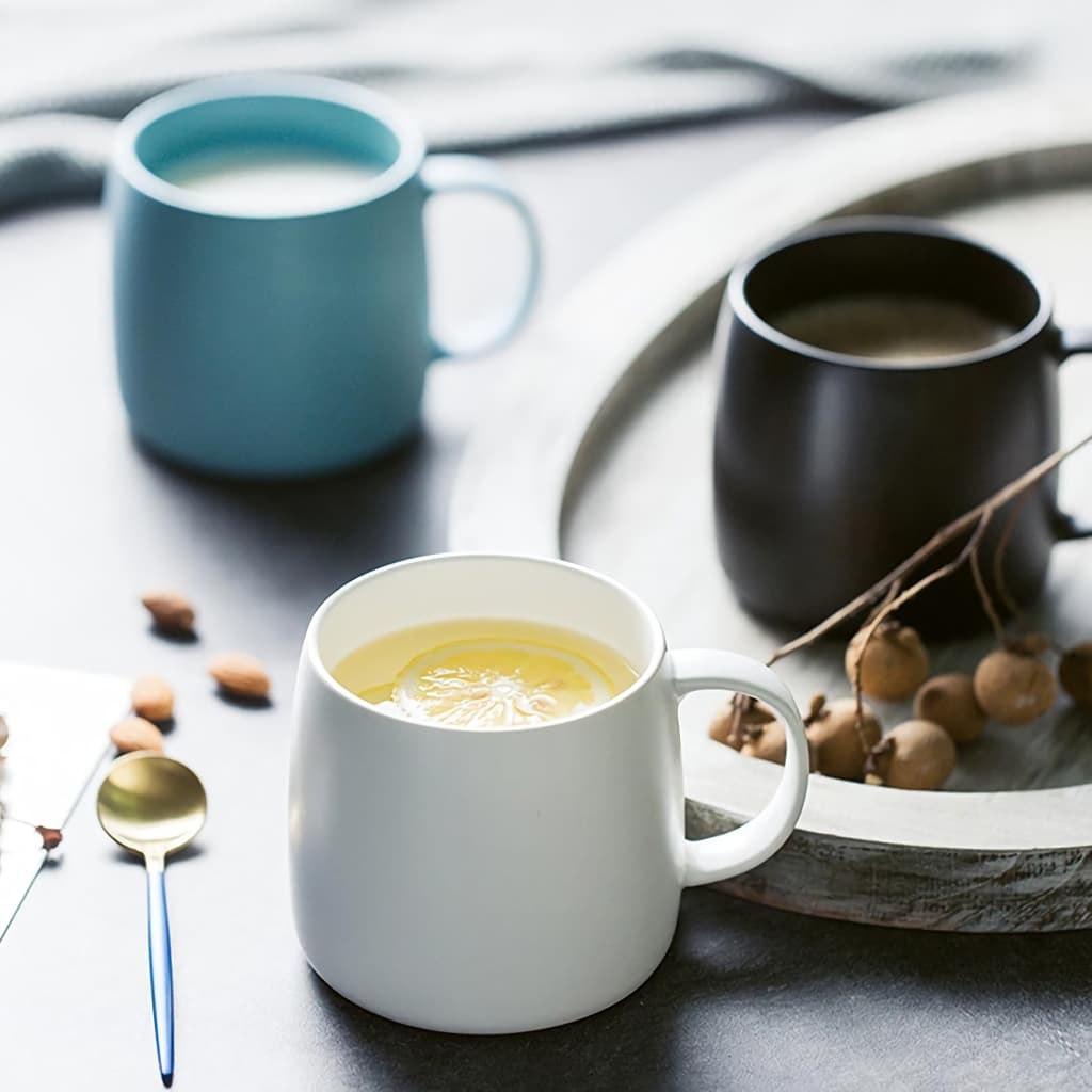 Three ceramic mugs sit on a dark surface: a white mug with a yellow liquid and lemon slice, a black mug with a dark beverage, and a blue mug with a pale drink. Nearby are a spoon, almonds, and decorative twigs on a round wooden tray.