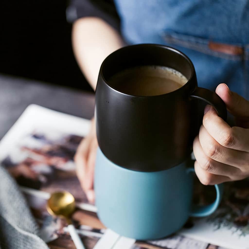 A person is holding a black mug filled with coffee, stacked on top of a blue mug. A golden teaspoon rests on an open magazine, and a gray cloth is nearby, creating a cozy and relaxed atmosphere.