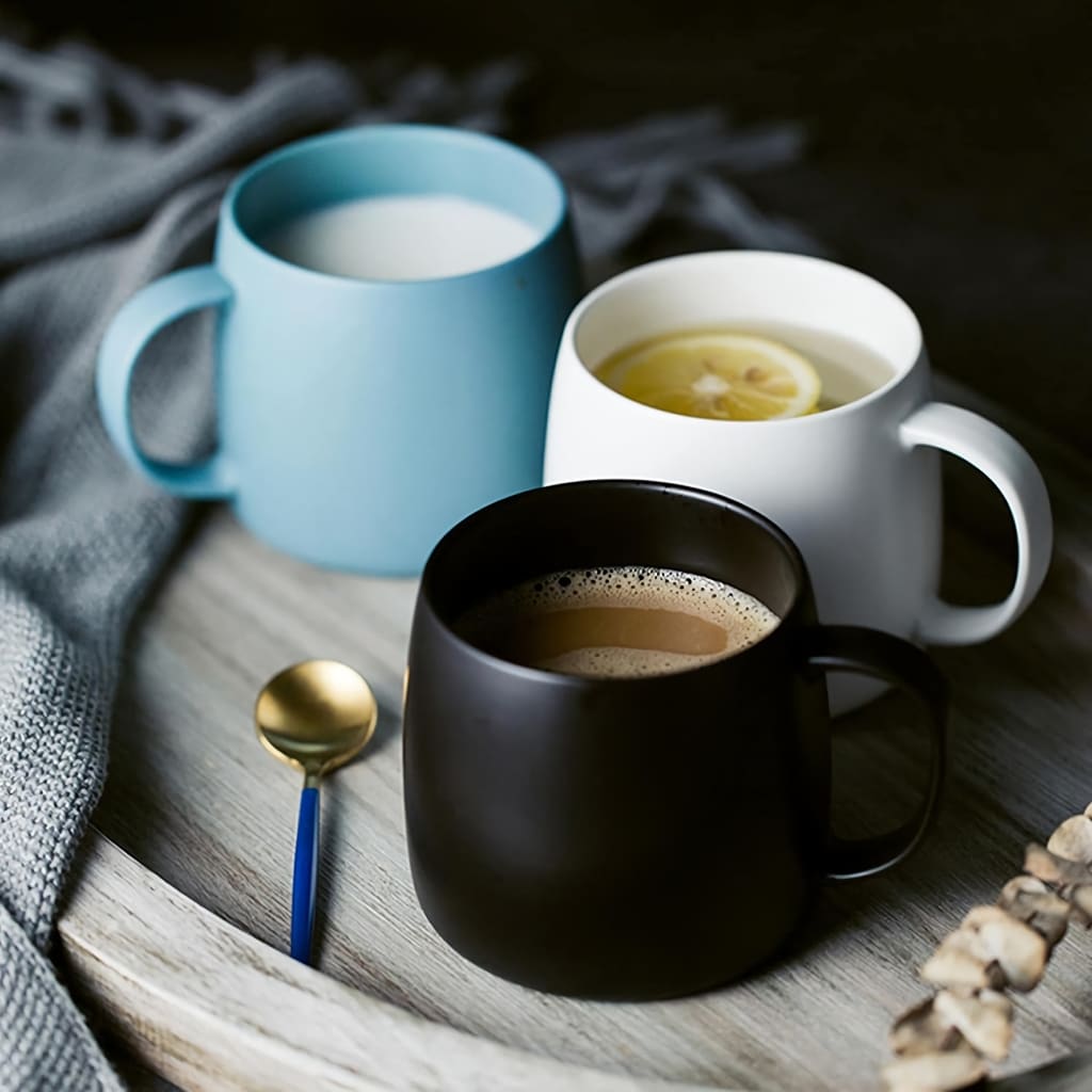 Three mugs on a wooden tray: a blue mug with milk, a white mug with lemon tea, and a black mug with coffee. A gold spoon with a blue handle lies beside them, next to a gray cloth.