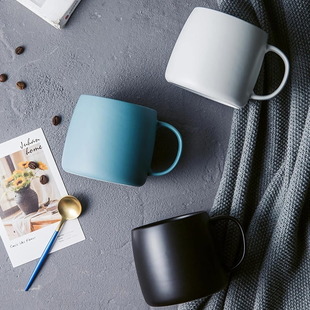 Three ceramic mugs in white, blue, and black are arranged on a textured gray surface. Nearby are a modern gold and blue spoon, scattered coffee beans, and a postcard with a vase of sunflowers. A cozy gray knit blanket completes the scene.