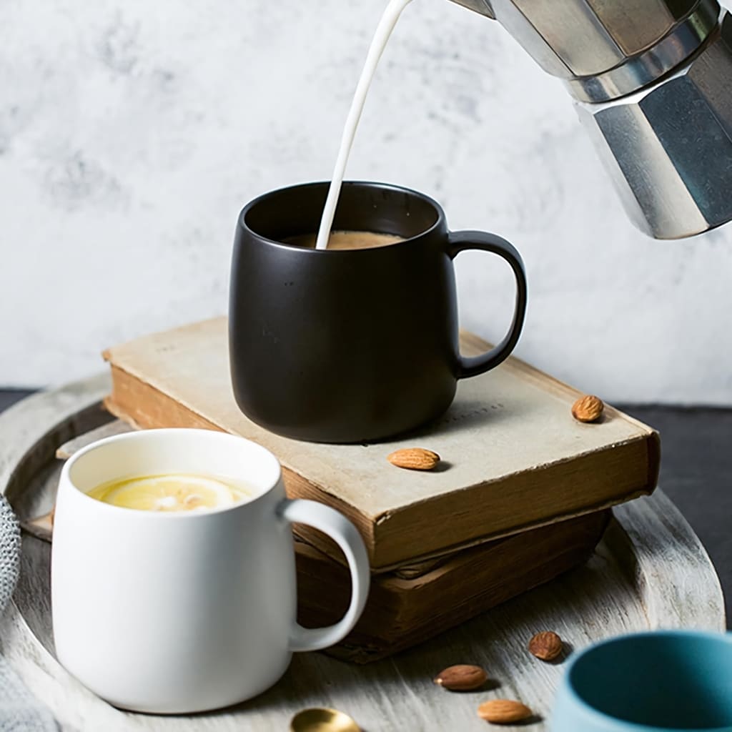 The image shows milk being poured into a black mug on top of stacked books. Next to it, there is a white mug on a wooden tray with scattered almonds. A moka pot is partially visible, suggesting a cozy coffee setup.