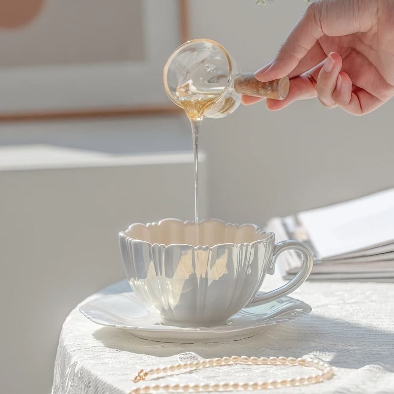 A hand pours honey from a small glass jar into a white teacup on a saucer. The scene is set on a sunlit tablecloth, with a pearl necklace beside the cup and soft, warm lighting highlighting the serene setting.