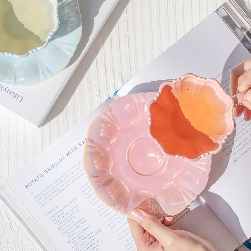 A person is holding an orange liquid-filled pink flower-shaped bowl, placing it on a matching pink plate. The plate is set on an open book with text about potato gnocchi. Sunlight casts soft shadows on the textured surface.