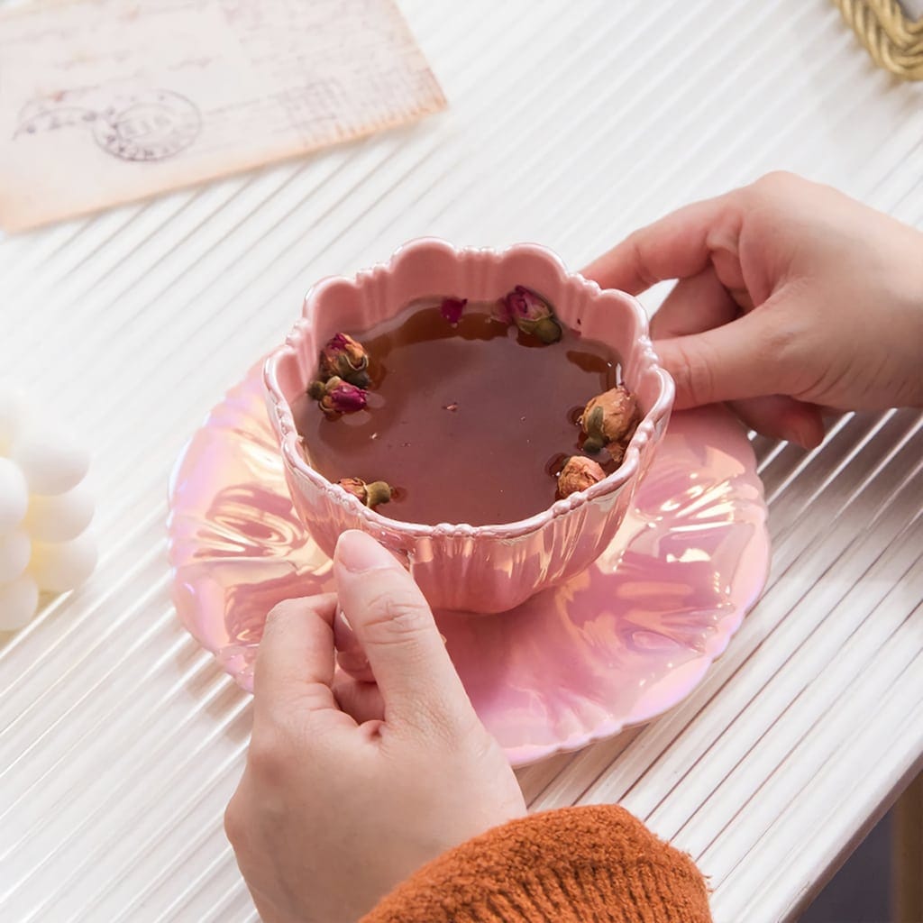 A person holds a pink, scalloped teacup filled with tea and rosebuds on a matching saucer. A textured white surface and a partially visible envelope are seen in the background.