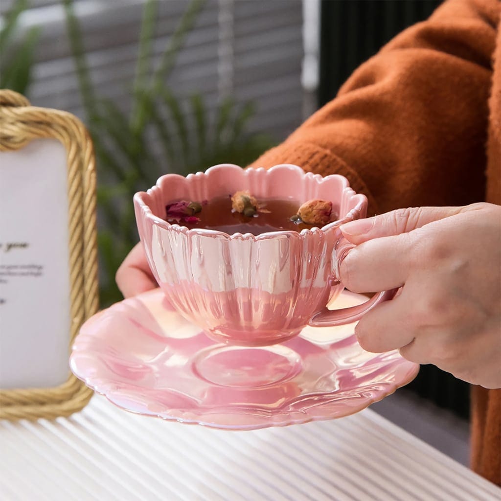 A person holding a pink, flower-shaped teacup and saucer filled with herbal tea, garnished with dried rosebuds. The background is blurred, with a glimpse of greenery and a framed sign.