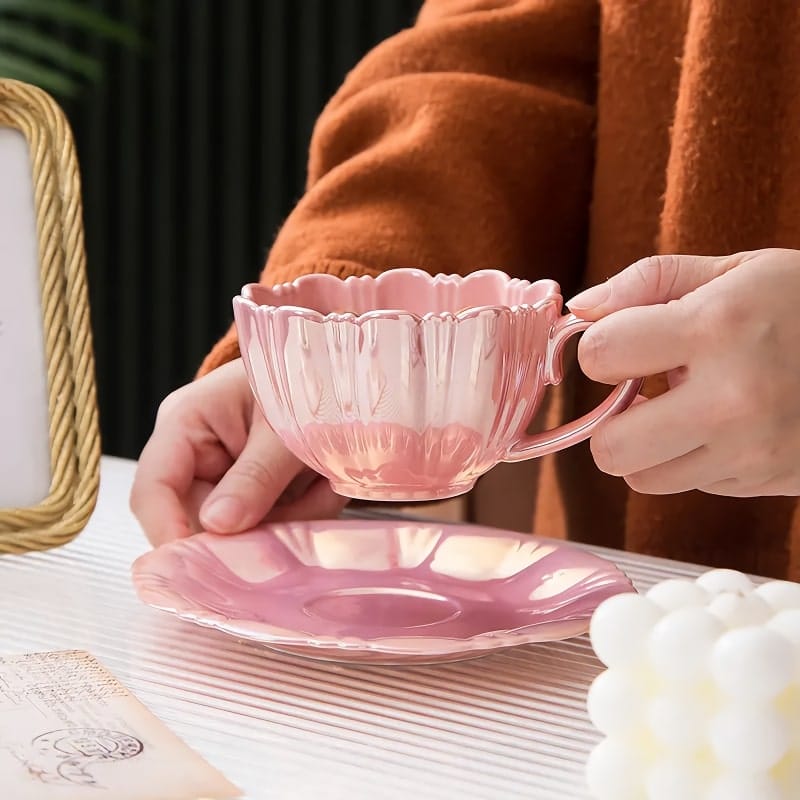 A person holding a pink, scalloped teacup above a matching saucer on a white striped surface. The person is wearing an orange sweater. Theres a picture frame and decorative items nearby.