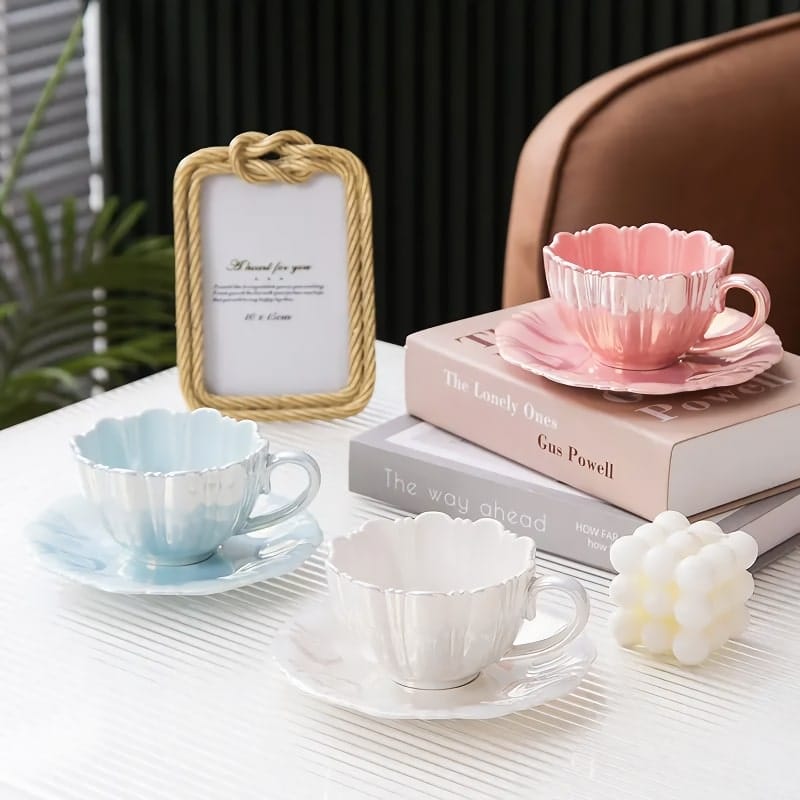 A stylish table setting with decorative teacups and saucers in pastel pink, white, and blue. They rest on a stack of books beside a rope-framed photo and a white bubble candle. In the background is a brown couch and vertical blinds.