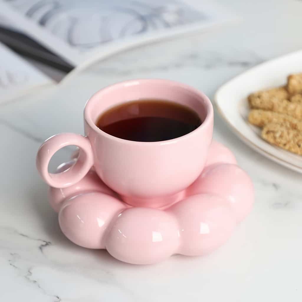 A pink teacup filled with coffee sits on a matching ring-shaped saucer. The cup and saucer have a glossy finish. In the background, theres an open magazine and a white plate with rectangular cookies on a marble surface.