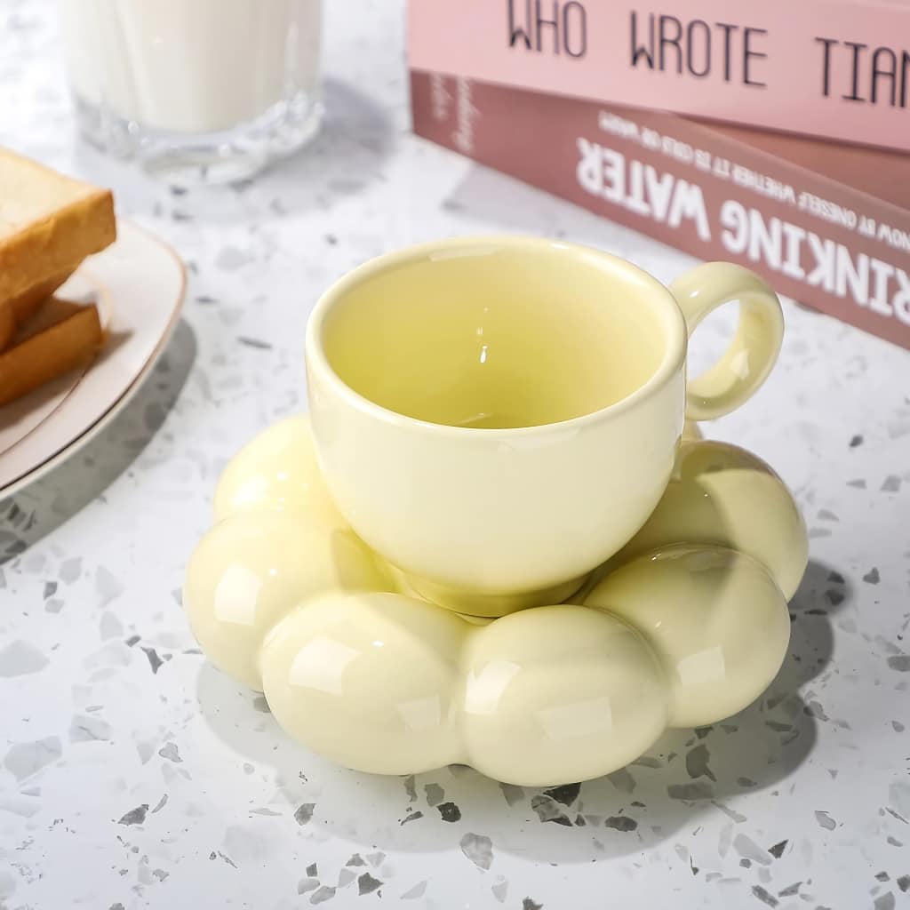 A yellow teacup sits on a matching cloud-shaped saucer. Nearby, there are slices of toast on a plate, a glass of milk, and books with pink covers resting on a speckled counter.