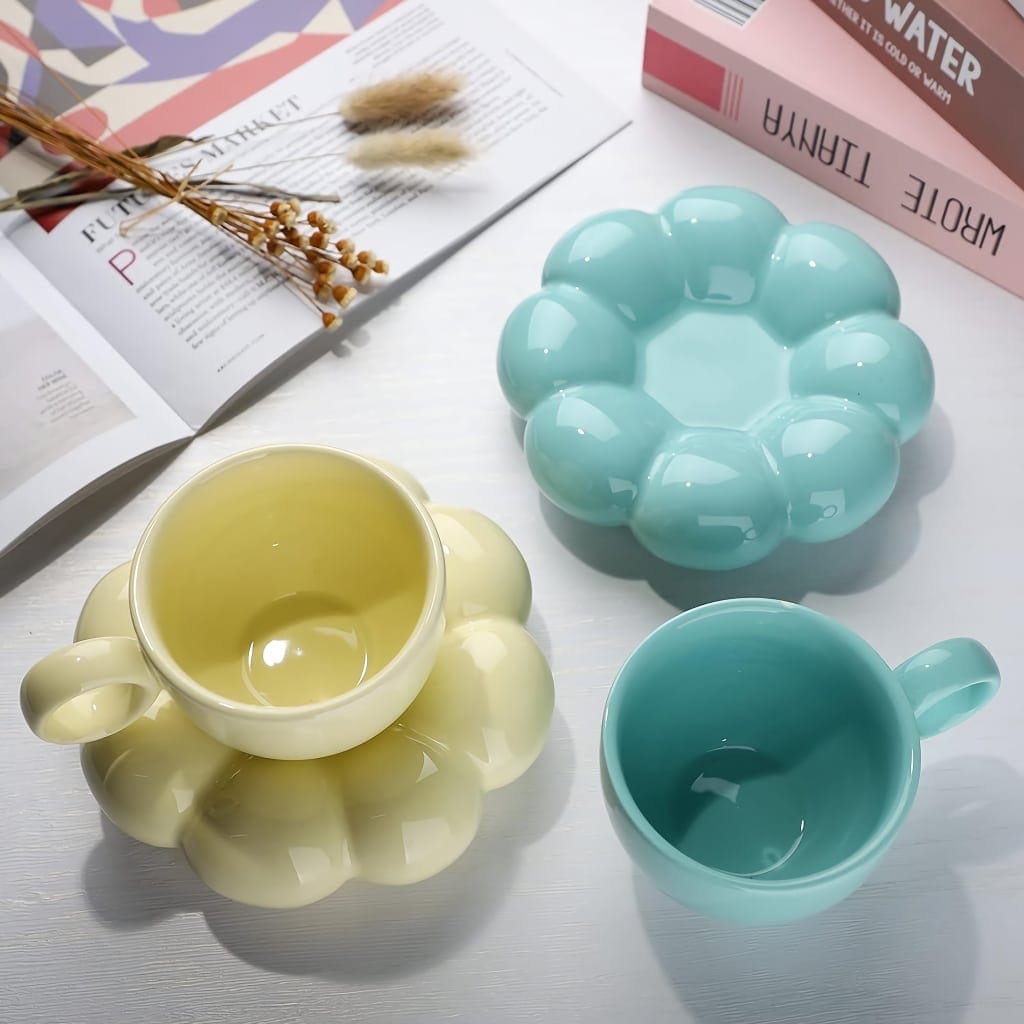 Pastel-colored ceramic cups and matching saucers with a bubble design are displayed on a white surface. An open magazine and decorative dried flowers are in the background.