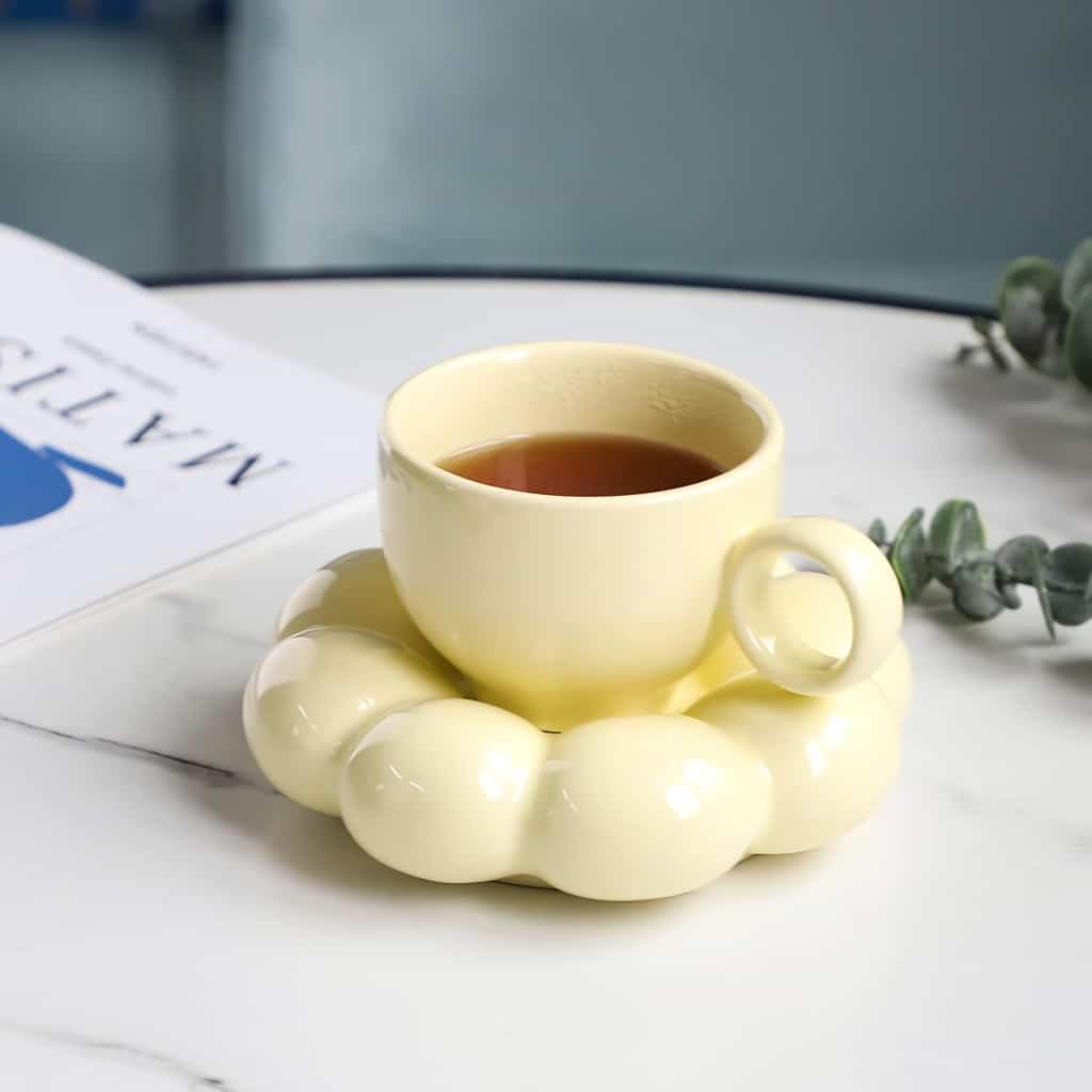 A yellow teacup filled with tea rests on a unique, round bubble-shaped coaster on a marble table. A magazine with visible text sits nearby, along with a sprig of eucalyptus.