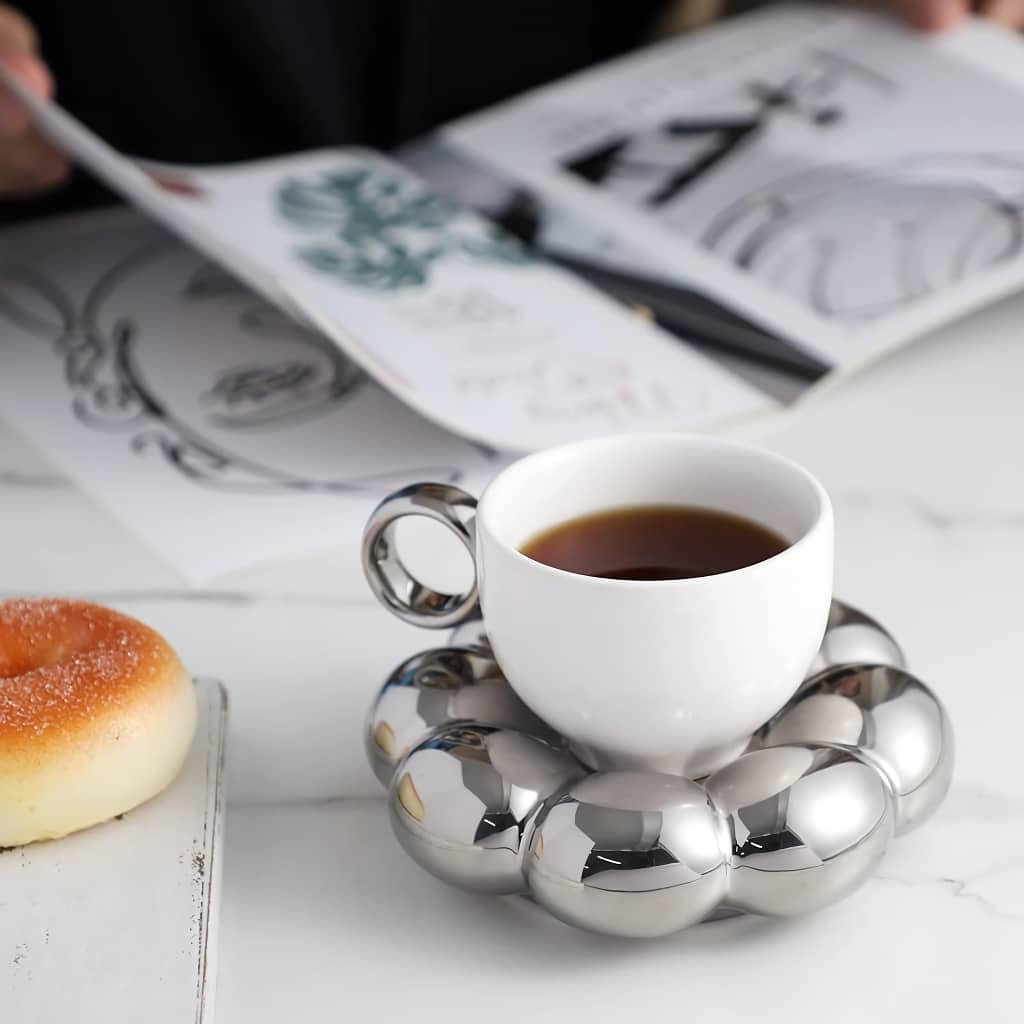 A white teacup filled with black coffee sits on a shiny, metallic ring-shaped saucer. Beside it on the table is a small pastry, and a person in the background is browsing through a book or magazine with illustrations.