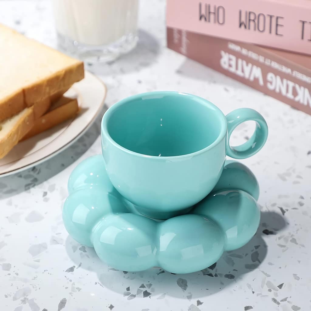 A teal ceramic teacup sits atop a matching, cloud-shaped saucer on a speckled surface. Nearby, a plate with bread slices and stacked books is visible, one titled WHO WROTE THIS. A glass of milk is also present.