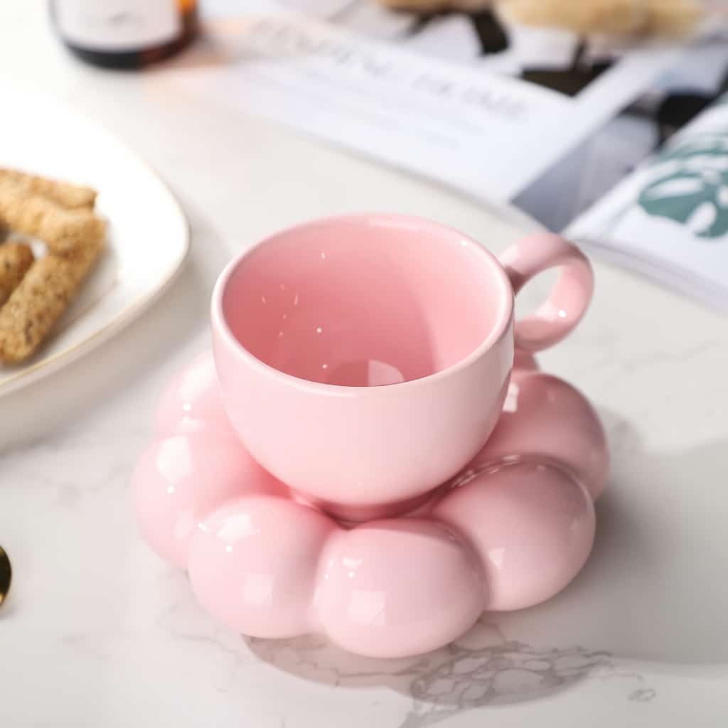 A pink teacup rests on a matching bubble-shaped saucer, placed on a marble countertop. Nearby, part of a white plate with breadsticks and a magazine are visible in the background.