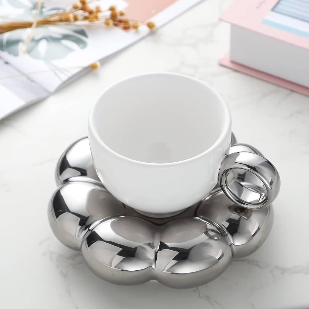 A white teacup with a metallic handle rests on a unique shiny silver bubble-shaped saucer. The background includes blurred elements like a book, a plant, and decorative dried flowers on a marble surface.