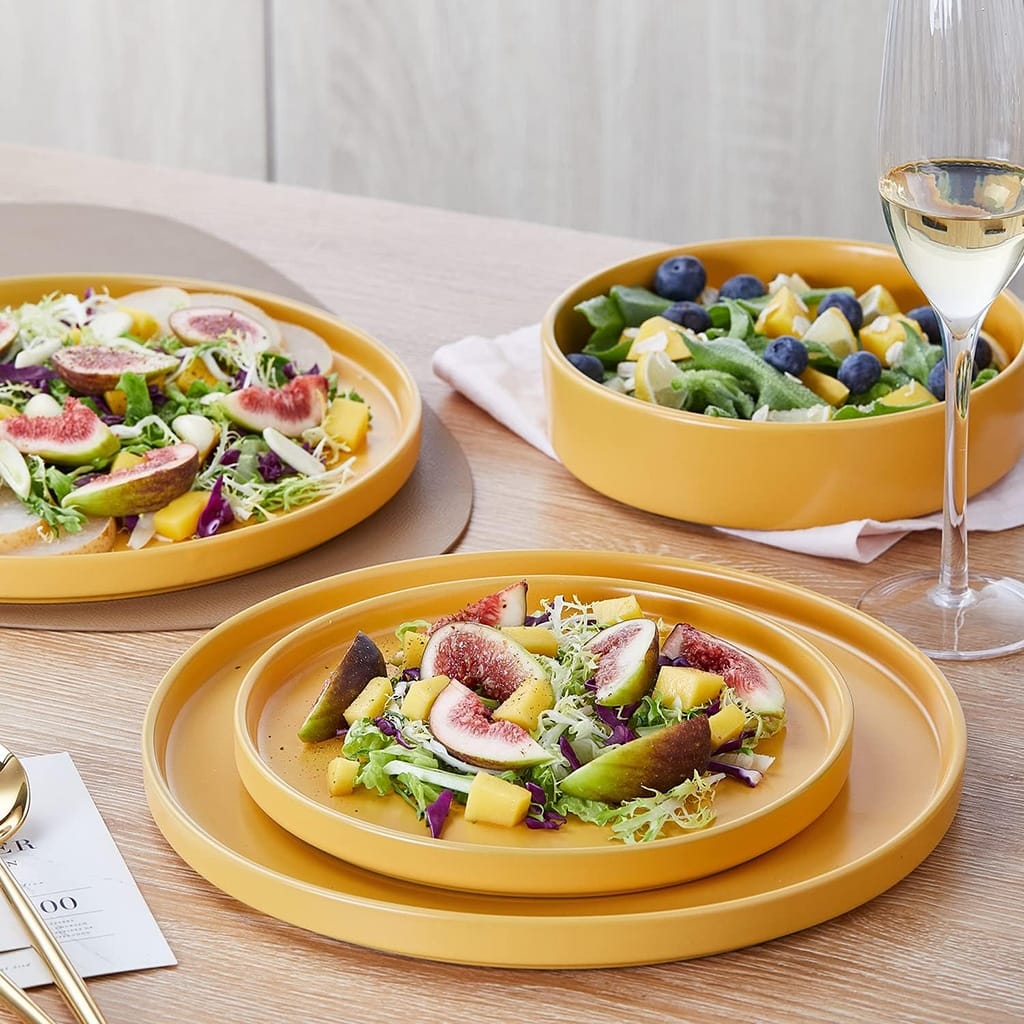 A stylish dining setup featuring a vibrant salad with figs, avocado, and mango on mustard-colored plates. A bowl of salad with greens and blueberries is on the side, accompanied by a glass of white wine on a wooden table.