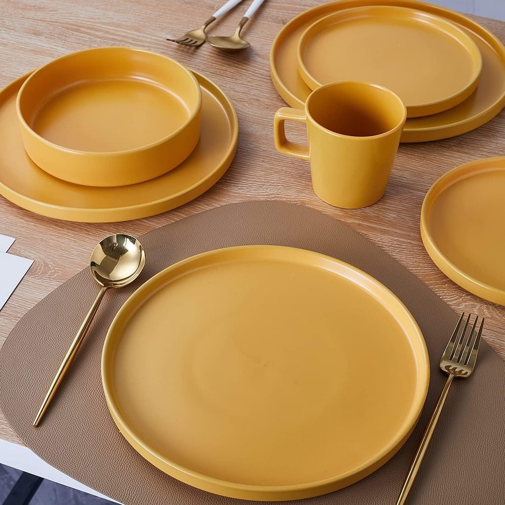 A table setting with mustard yellow dishes, featuring plates, bowls, and a mug on a wooden table. Gold cutlery and a brown placemat are also present, creating a cohesive and elegant look.