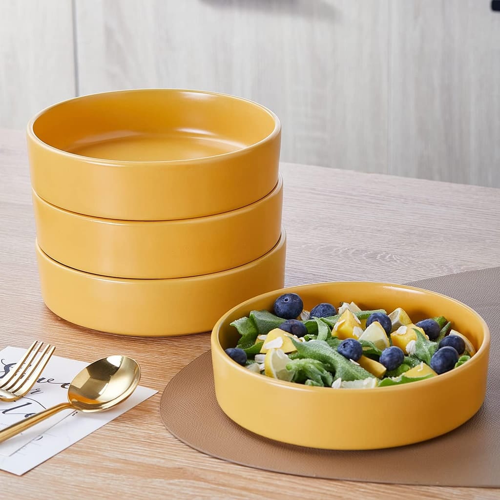 A stack of four yellow ceramic bowls next to a filled bowl with a salad of kiwi, blueberries, and diced fruit. A gold fork and spoon are placed nearby on a napkin. The setup is on a light brown placemat on a table.