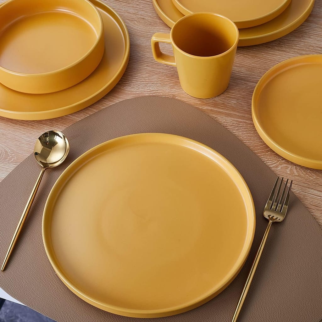 A table setting featuring yellow plates, bowls, and a mug on a wooden surface with a brown placemat. A gold spoon and fork are placed beside a large plate, creating a warm and elegant dining arrangement.