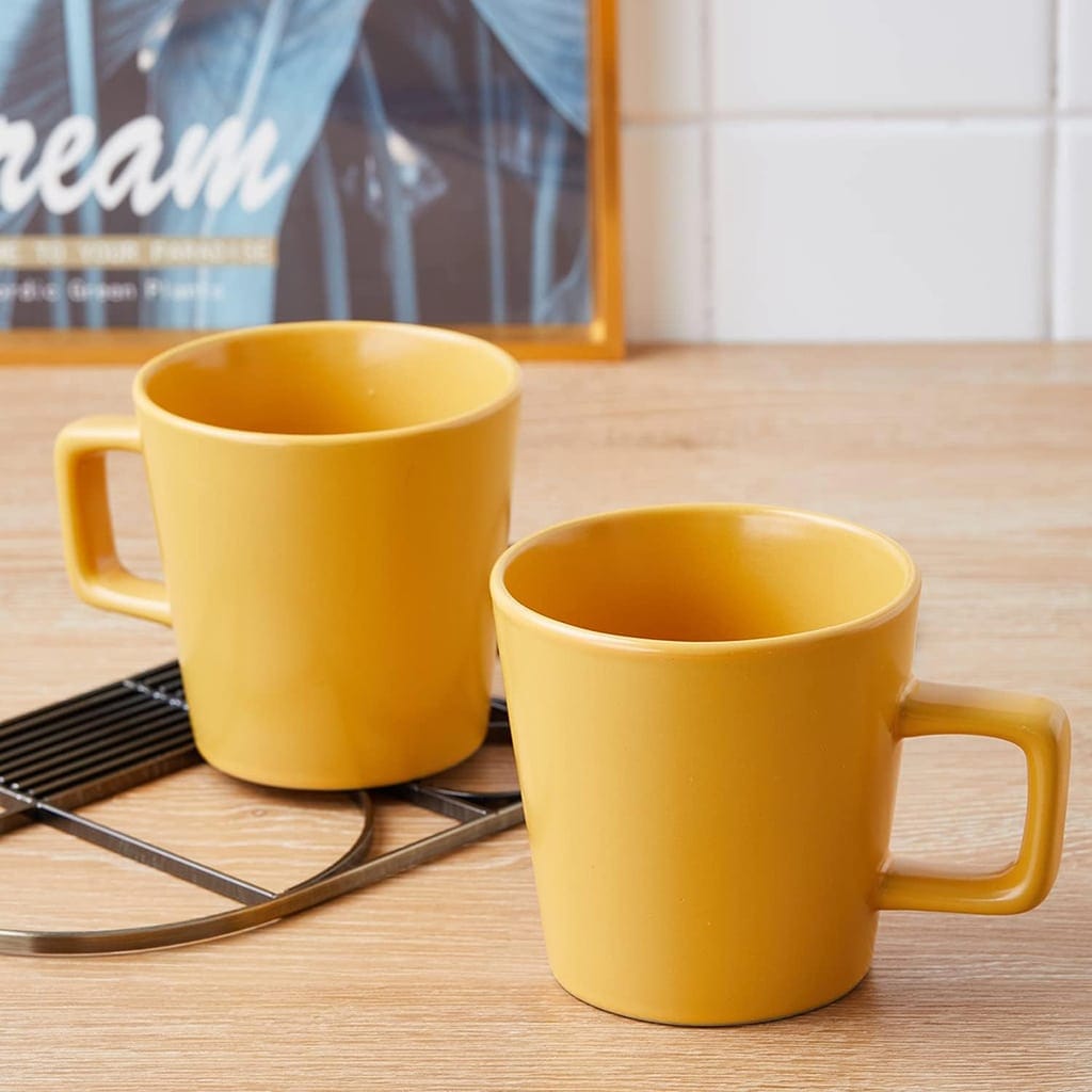 Two yellow mugs with square handles sit on a wooden countertop. One mug is on a black metal trivet. A partially visible framed picture and white tile backsplash are in the background.