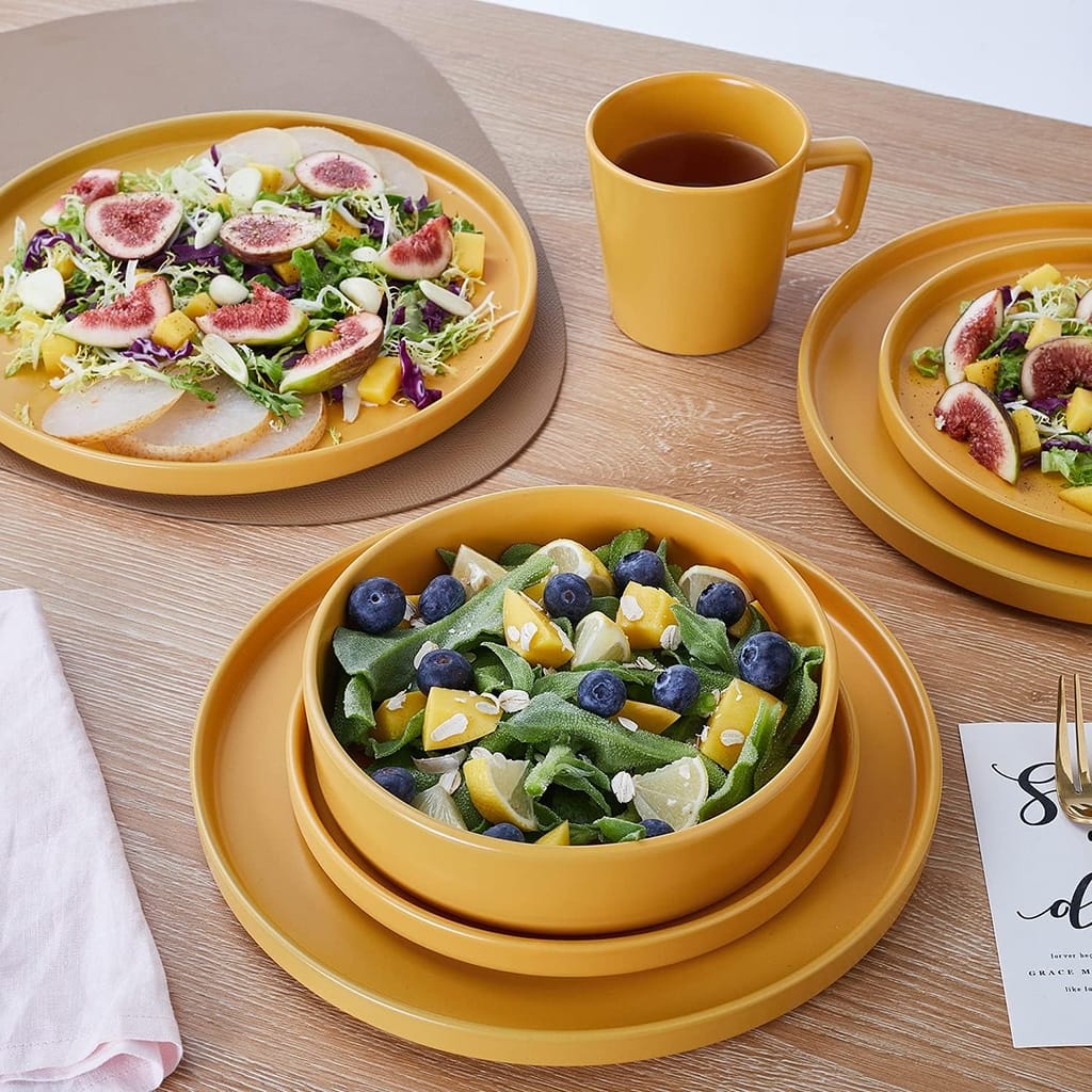 Two orange plates with vibrant salads featuring figs, blueberries, and citrus segments are displayed on a wooden table with a cup and two matching bowls. A pink napkin is folded beside the setting.