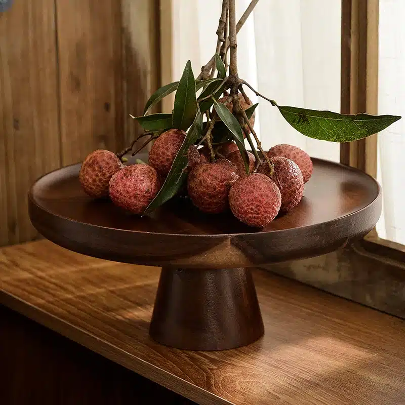 A bundle of lychees with leaves placed on a round, dark wooden pedestal stand with a wooden backdrop and soft natural light filtering through a nearby window.