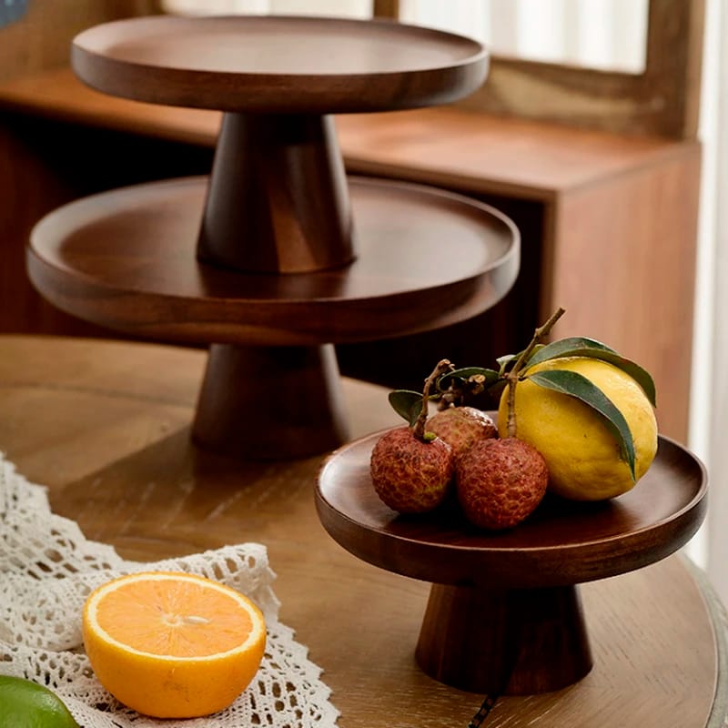 Wooden cake stands of varying heights are displayed on a table. The smallest stand holds a lemon and two lychees. An orange half is on the table, which is decorated with a lace cloth. A blurred background suggests a cozy indoor setting.