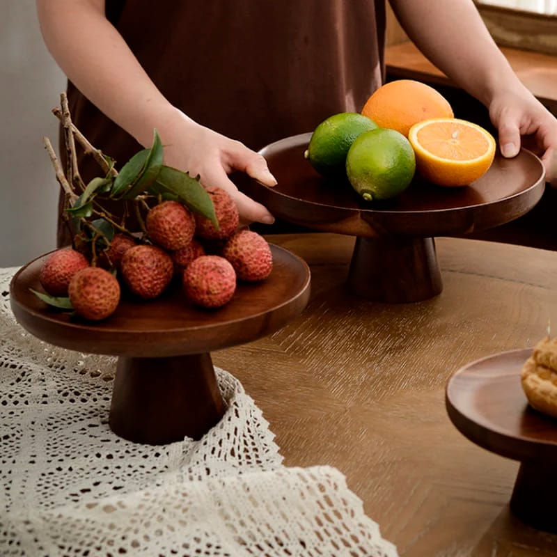 A person arranges wooden cake stands on a table. One stand holds lychees and leafy branches, while the other holds citrus fruits like oranges and limes. The table is covered with a white lace table runner.