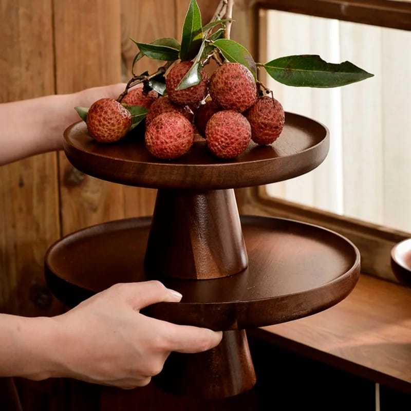 A person arranging lychees with leaves on a two-tiered wooden stand near a window. The stand has a polished, dark finish and contrasts with the rustic wooden background.