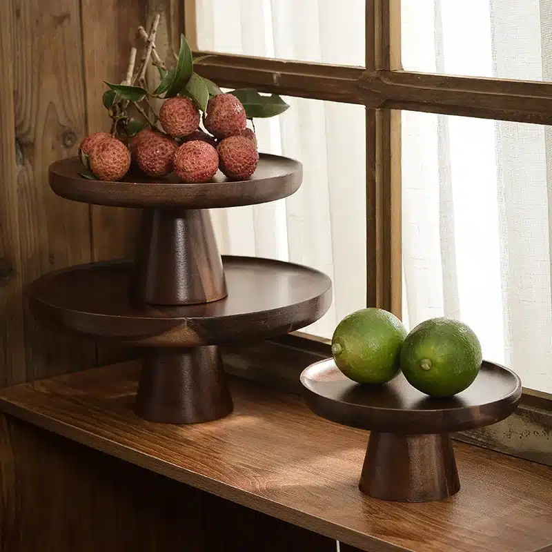 Two wooden tiered stands by a window hold fruit. The taller stand has lychees with leaves, while the shorter one has two limes. Both stands and fruit are set on a wooden surface next to a window with sheer curtains.