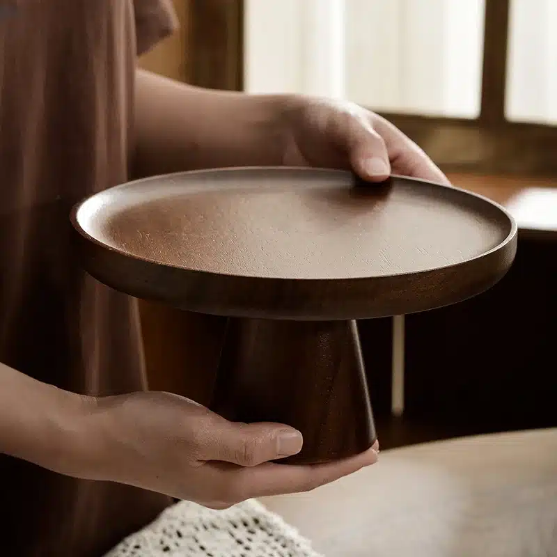 A person holding a round, wooden cake stand with a pedestal base, set in a softly lit room. The background shows a window with beige curtains, creating a warm and cozy ambiance.