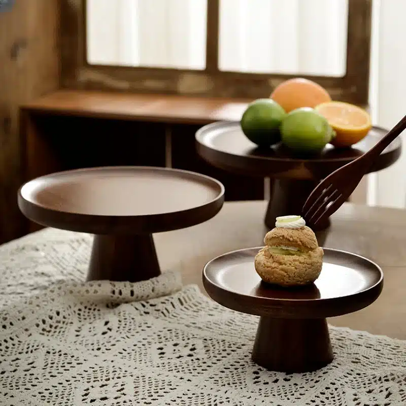 Wooden cake stands on a table with a lace tablecloth. One stand holds a pastry with cream, while another holds citrus fruits. A fork is reaching for the pastry. A window with curtains is in the background.